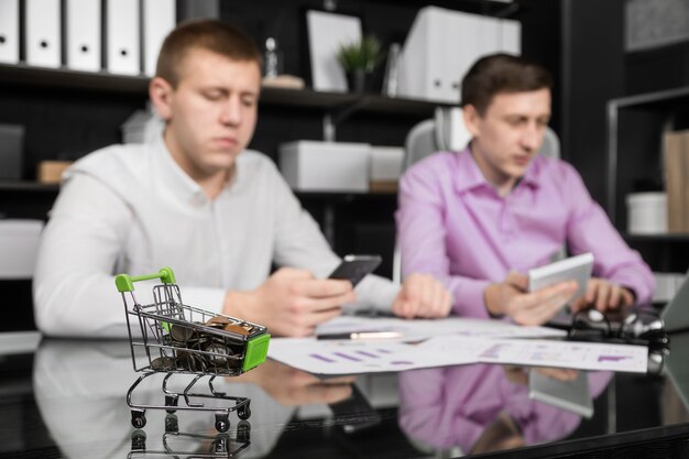 Dummy shopping cart with coins on the office table on the of two businessmen