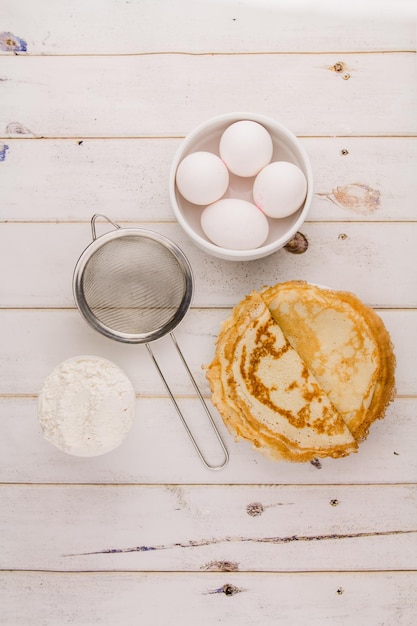 Dunne heerlijke ronde pannenkoeken op een witte plaat op een houten tafel in de keuken met eieren en bloem