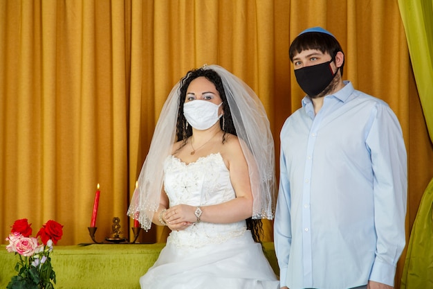 During the chupa ceremony in the synagogue, the masked jewish\
bride and groom stand side by side. horizontal photo