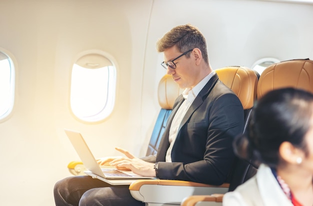 Durante un volo un uomo d'affari in abiti formali e occhiali guarda fuori dal finestrino dell'aereo mentre lavora su un computer portatile servizio con accesso a internet a bordo e un concetto di viaggio d'affari