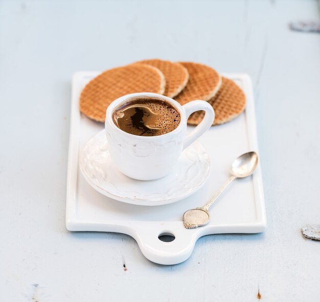 Dutch caramel stroopwafels and cup of black coffee on white ceramic serving board over light blue wooden backdrop