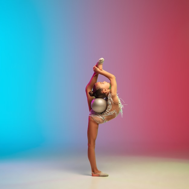 Dynamic. Little caucasian girl, rhytmic gymnast training, performing isolated on gradient blue-red studio background in neon. Graceful and flexible, strong child. Concept of sport, motion, action.