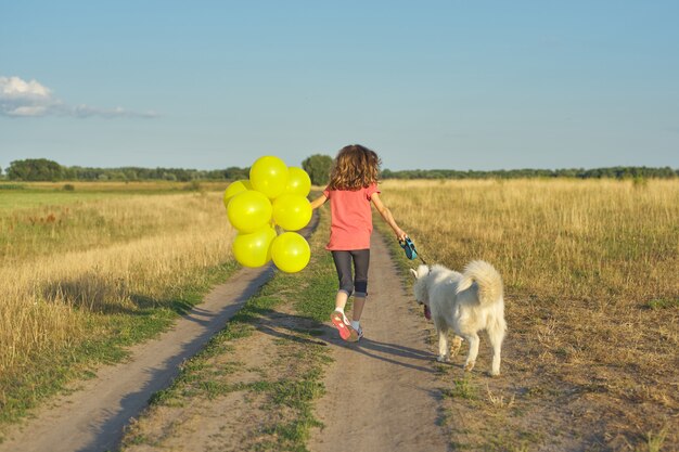 Dynamic outdoor portrait of running girl with white dog and yellow balloons