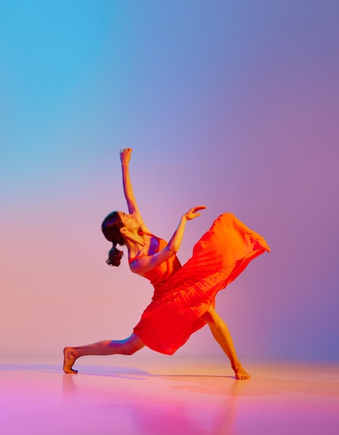 Dynamisch beeld van artistieke jonge vrouw dansen in elegante rode jurk tegen veelkleurige kleurovergang