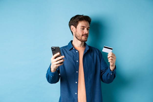 E-commerce concept. Man paying for online payment, reading credit card number and holding mobile phone, standing on blue background.