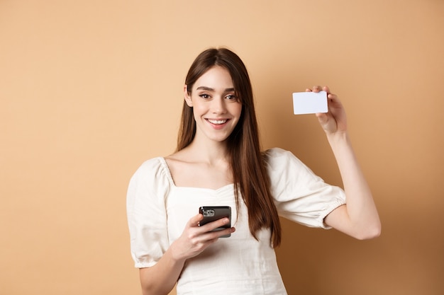 Концепция электронной коммерции. Улыбающаяся женщина показывает пластиковую кредитную карту во время покупок в Интернете на мобильном телефоне, стоя на бежевом фоне.