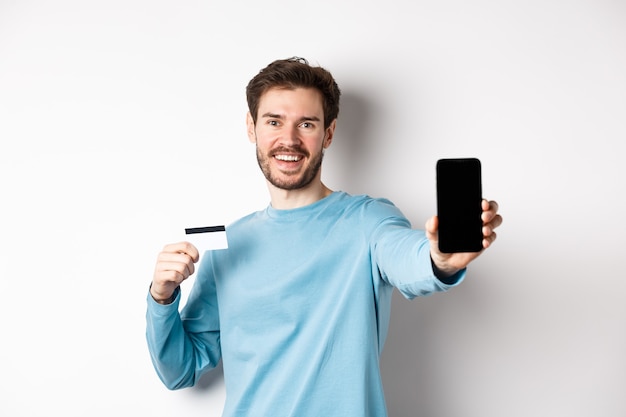 E-commerce en winkelconcept. Glimlachende blanke man met plastic creditcard en leeg smartphonescherm, online app, witte achtergrond aanbevelen.