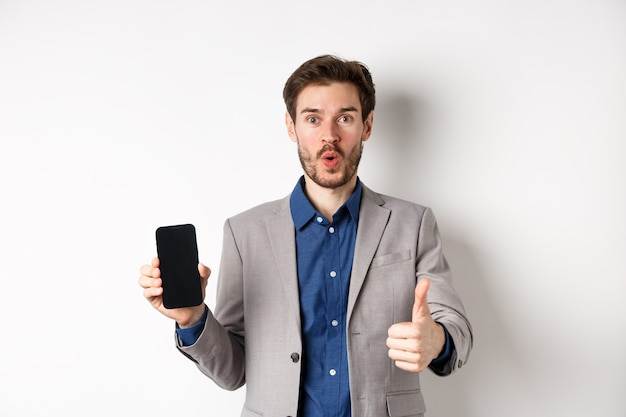 Концепция электронной коммерции и покупок в Интернете. Взволнованный бизнесмен рекомендует финансовое приложение, показывая пустой экран смартфона и большой палец вверх, белый фон.