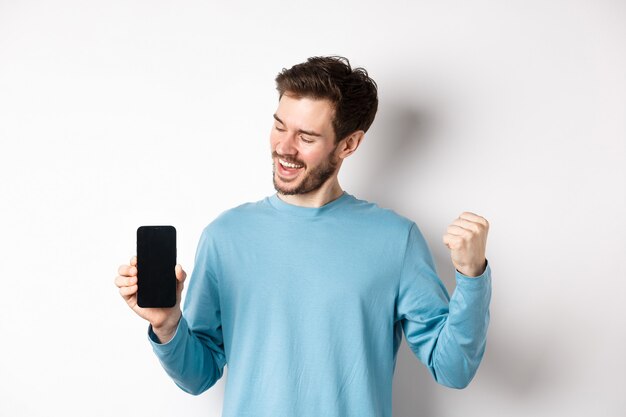 E-commerce and shopping concept. Lucky man winning online, showing empty smartphone screen and triumphing, saying yes with fist pump, standing over white background