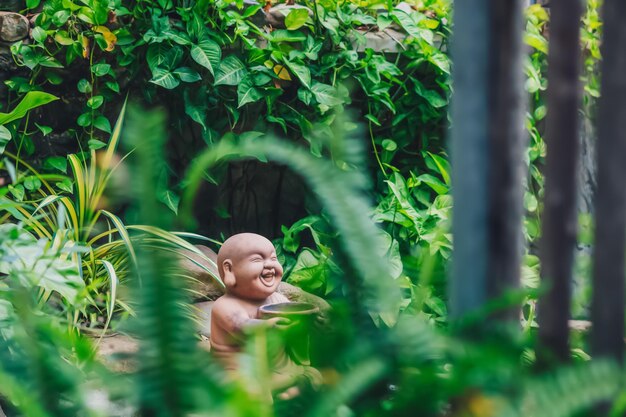 E statuette figurine symbol brown stone buddha smile hold bowl
copy space nature tropical leaves plants garden mysterious calm
soul meditation mood buddhist culture bright green more tone in
stock