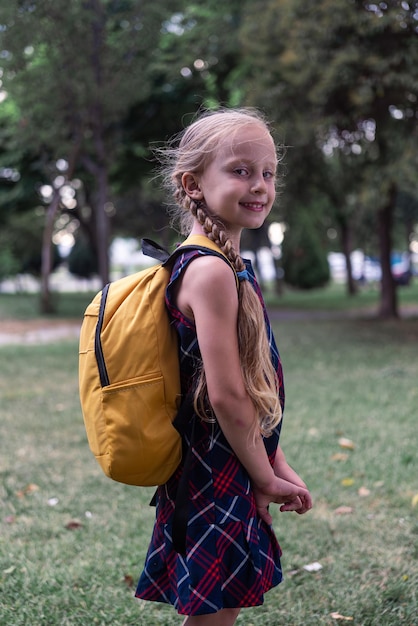 Photo eady for school blonde schoolgirl with yellow backpack beams with joy portrait in city park