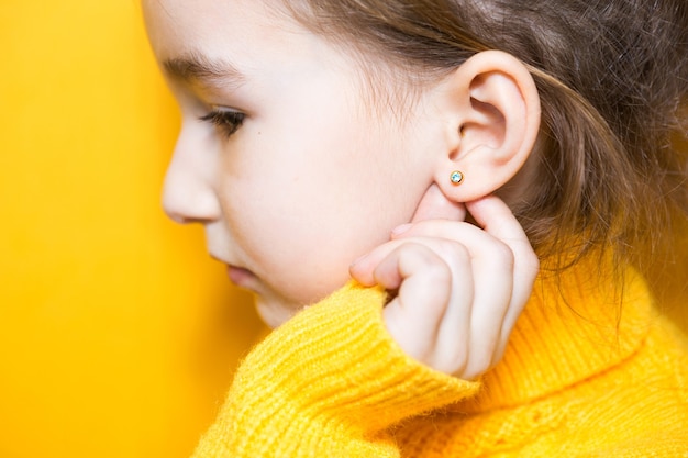 Ear piercing in a child - a girl shows an earring in her ear made of a medical alloy. Yellow background, portrait of a girl in profile.