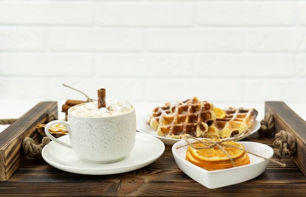 Early breakfast coffee with marshmallows and a stick of cinnamon and Belgian waffles lie on a wooden tray