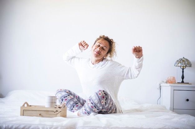 Early morning wake up concept with happy cheerful caucasian beautiful adult woman stretching and smiling - white colors and light in the bedroom athome or hotel - stay home quarantine