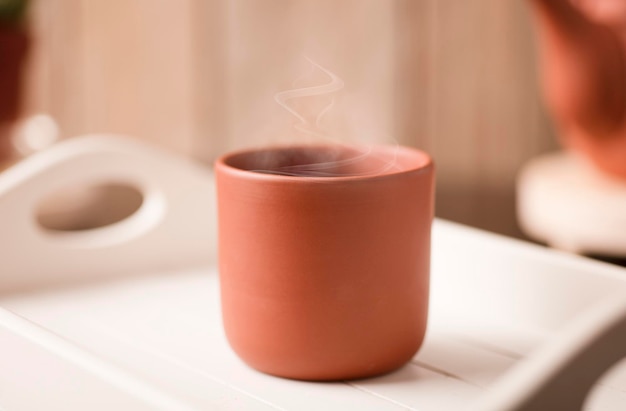 Earthenware mug with hot steam coffee or tea on a white wooden tray with partial view of the kettle