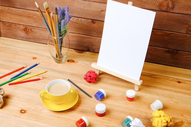 Easel, art brushes and coffee on a wooden table. Workplace of the artist. Creativity and hobby concept.