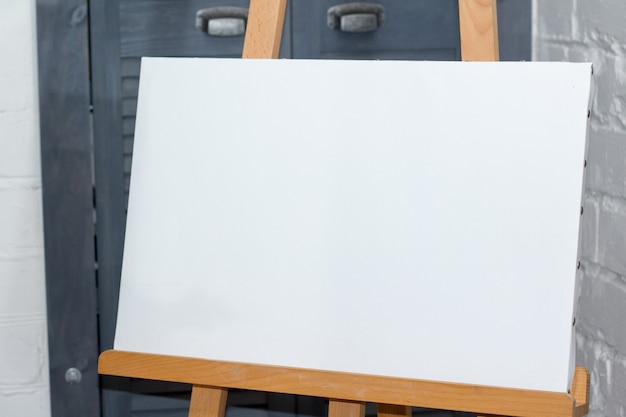 An easel with a clean white canvas is ready for work against a white brick wall