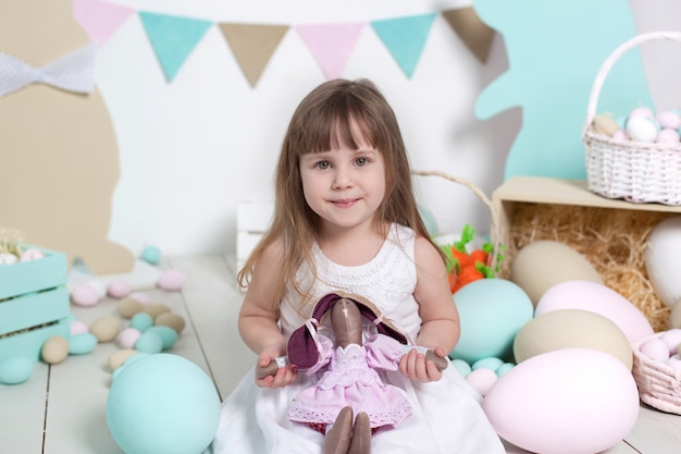 イースター！白いドレスの美しい少女は、おもちゃのウサギを抱擁します。子供は贈り物を受け取り、休日を楽しんでいます。多くの異なるイースターエッグ、カラフルなイースターインテリア。イースターのウサギ。春の装飾