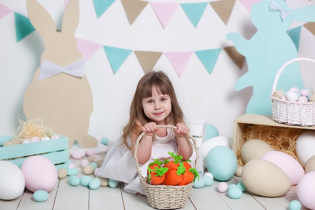 Easter! A beautiful little girl in a white dress is sitting with an Easter basket and a carrot. Rabbit, colorful Easter eggs. Easter interior. Family celebration. Agriculture. Child and garden.