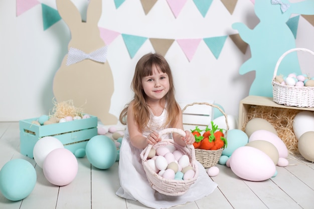 Easter! Beautiful little girl in a white dress with a basket with multi-colored Easter eggs and Easter bunnies. Easter decoration, spring decor. Family holidays, traditions. Little farmer. Harvesting