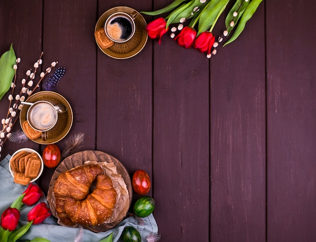 Photo easter breakfast with aromatic fresh espresso, croissants, colored eggs, red tulips and willow. coffee with pastries, flowers on a wooden table. view from above. copy space