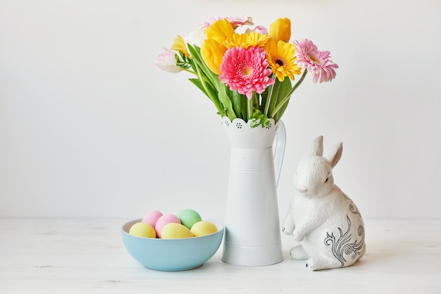 イースターのウサギとキッチンテーブルの上のイースターエッグ。チューリップと尾根とカラフルな卵の花束とテーブルの上に座っている白いウサギ。ウサギと卵のイースター装飾。イースターカードテンプレート
