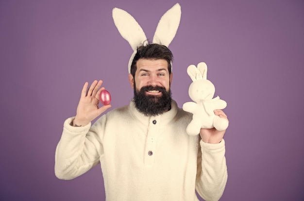 Easter bunny hunt begins Happy man with rabbit ears holding bunny toy and egg Bearded man in rabbit costume with easter egg and hare toy Spring new life and fertility Spring holiday celebration