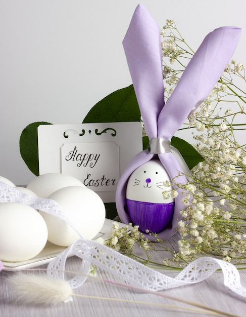 Фото Пасхальный кролик с пасхальными яйцами и цветами, с картой happy easter на белом деревянном столе