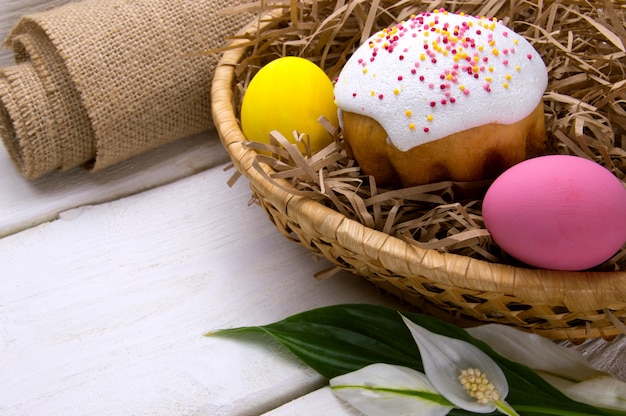 Easter cake and colored easter eggs in basket nest, sackcloth and flower on white wooden surface