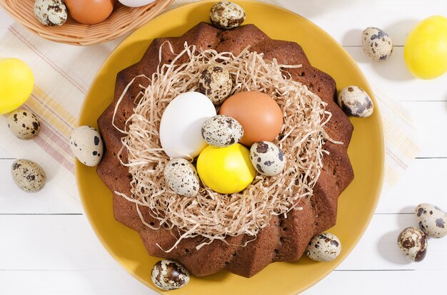 Easter cake with dyed eggs in a nest, daffodils in a cup on a white rustic wooden background