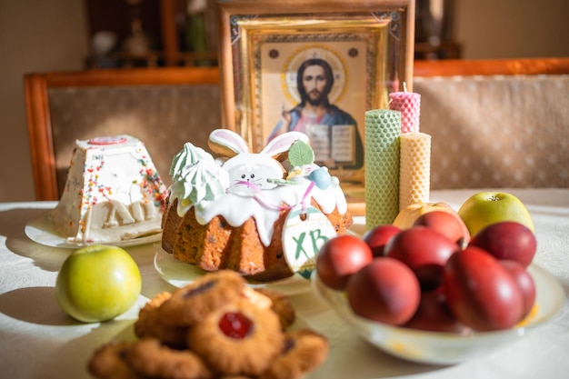 Пасхальный кулич с крашеными яйцами, яблоками и печеньем на столе на домашней кухне. Церковные иконы и свеча на фоне темы православной религии.