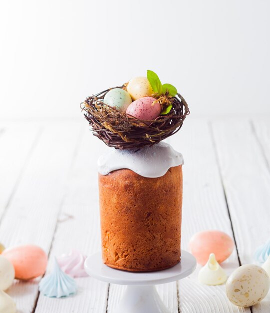 Easter cake with white icing stands on a white stand On the top of Easter cake is nest with painted eggs Next to on a wooden table different decorations