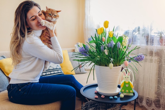 Празднование Пасхи дома. Счастливая женщина обнимает кошку, отдыхающую на диване. Весенние цветы в горшке, украшенные яйцами и кроликом на журнальном столике.