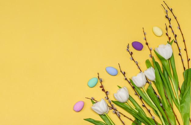 Easter colored eggs, a bouquet of white tulips and pussy-willow branches on a yellow background, copy space