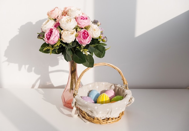 Easter colorful eggs in busket on white wooden table. Bouquet of rose flowers on table. Happy Easter. Springtime mood.