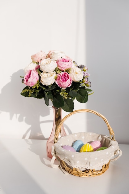 Easter colorful eggs in busket on white wooden table. Bouquet of rose flowers on table. Happy Easter. Springtime mood.