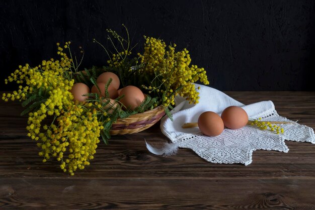 The easter composition Eggs a bouquet of yellow mimosa and church candles on wooden table closeup