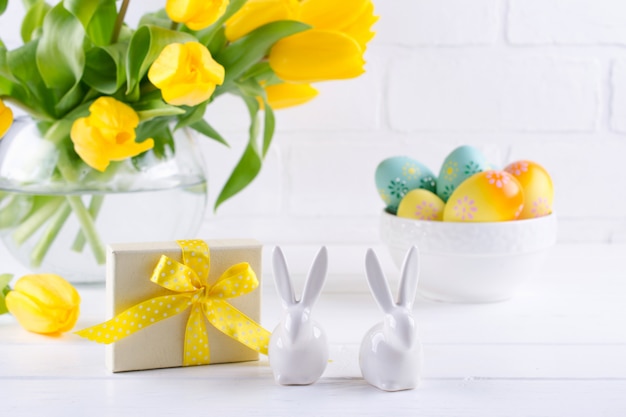 Easter composition with bouquet of yellow tulip flowers in glass vase and two white ceramic rabbits on white background