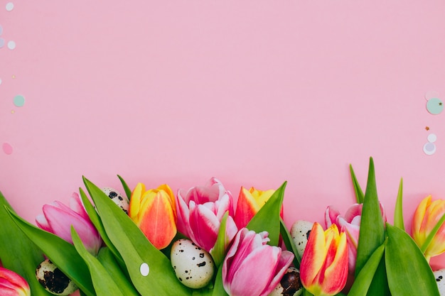 Easter concept, golden star decorations, vibrant confetti and multicolored tulips and quail eggs on pink background.