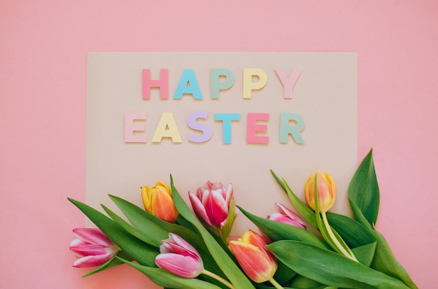 Easter concept. Happy easter paper cut inscription with pink and red tulips on pink background. Copy space, flat lay.