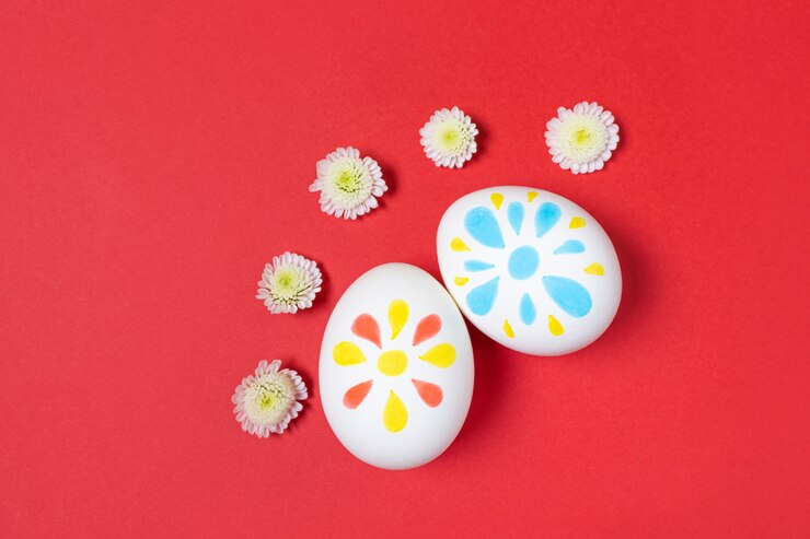 Easter concept. White eggs with marker drawings of petals. Manual coloring. Red, yellow and blue shades. Around - small white flowers. Flat lay, top view, copyspace.
