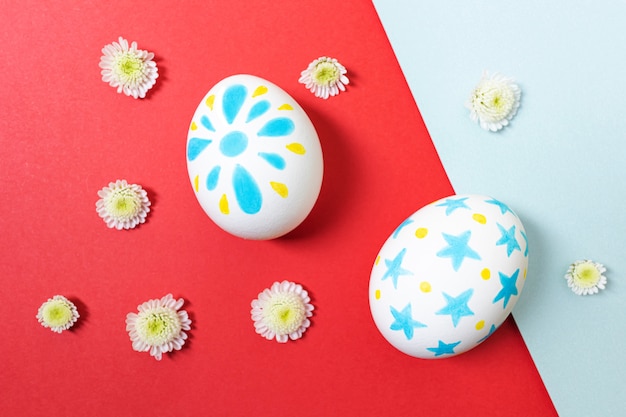 Easter concept. White eggs with marker drawings of petals and stars. Manual coloring. Yellow and blue shades. Nearby are small white flowers. Flat lay, top view, copyspace.