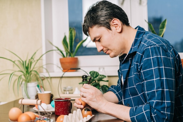 Пасхальный день обрезает руки взрослого мужчины, красящего яйца на деревянном фоне, сидящего на кухне с