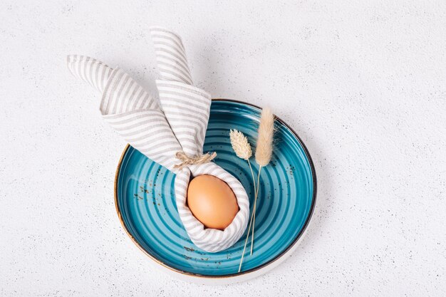 Easter egg decorated as a bunny in a blue plate on a white concrete background