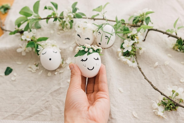 Easter egg with drawn cute face in floral wreath in person hand on background of eggs and petals