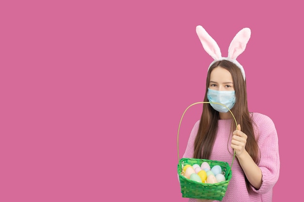 Easter eggs background holiday discounts and sales concept\
happy girl in bunny ears and medical mask with basket of decorated\
eggs smiling isolated on a pink background in studio