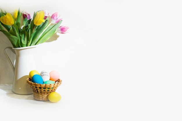 Easter eggs in basket and yellow tulips bouquet in vase near wall
