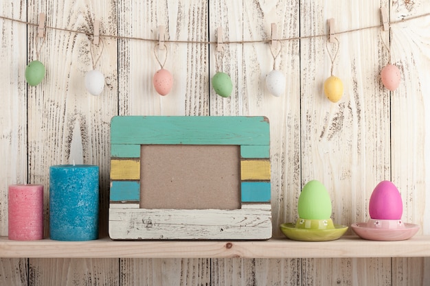 Easter eggs, blank frame and candles on wooden background