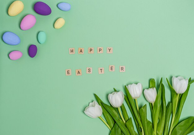 Easter eggs colored , a bouquet of white tulips and the word Happy Easter on a green table copy space, flat lay. Place for text. The concept of the holiday, spring. View from above.