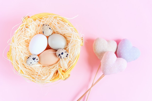 Uova di pasqua di diversi colori e uova di quaglia in un nido di paglia e cuori decorativi tessili su una superficie rosa, vista dall'alto, piatto laici. concetto di pasqua.